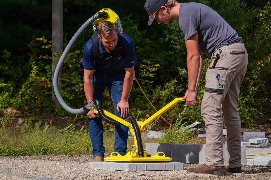 Two people using a yellow and black device on a concrete surface with greenery in the background